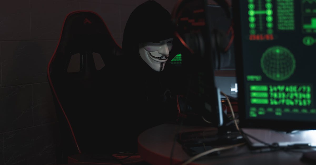 An anonymous hacker wearing a Guy Fawkes mask sits at a computer in a dimly lit room, engaged in cyber activities.